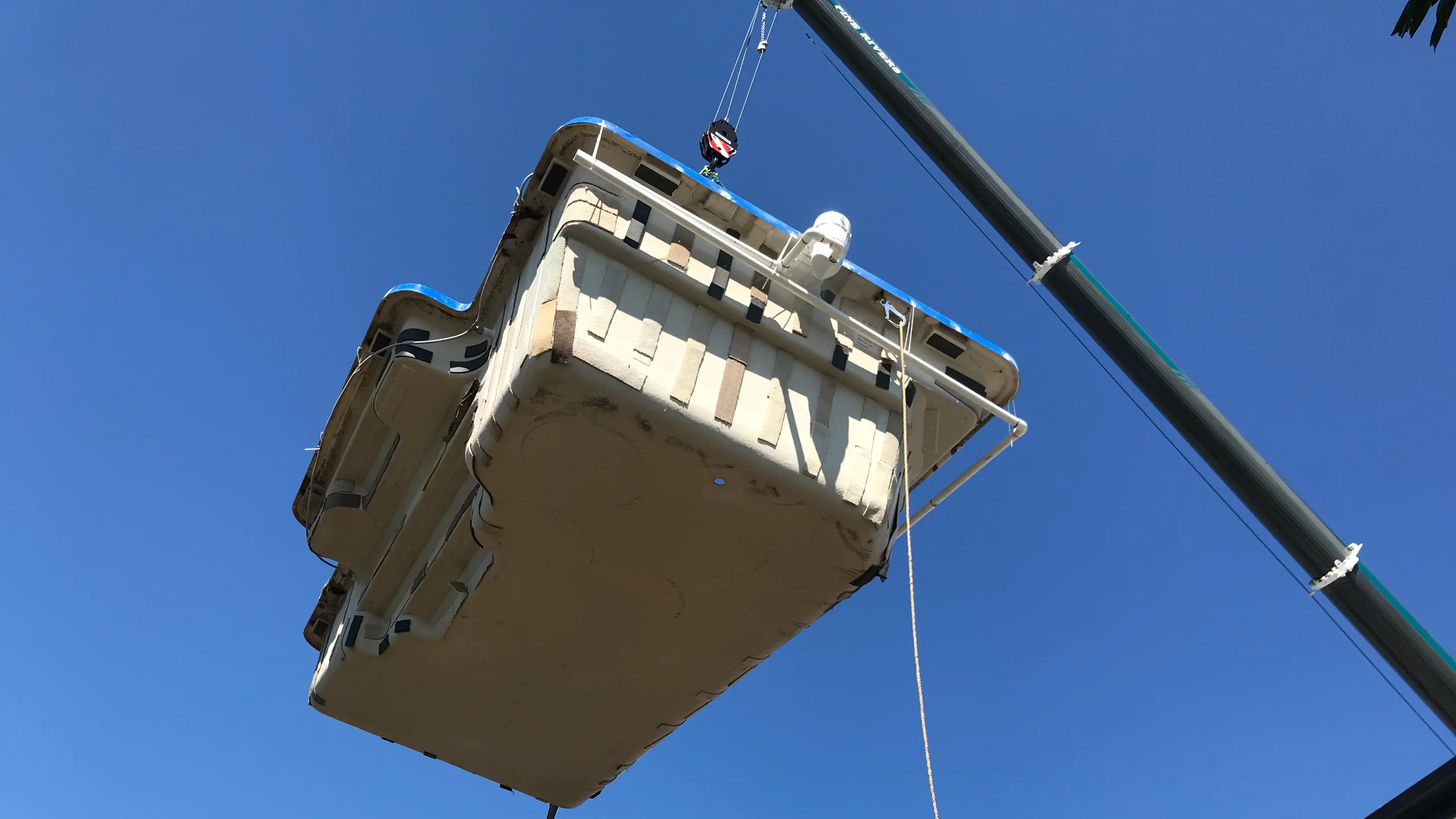 Fibreglass pool shell suspended from a crane hook against clear blue sky during a backyard lift