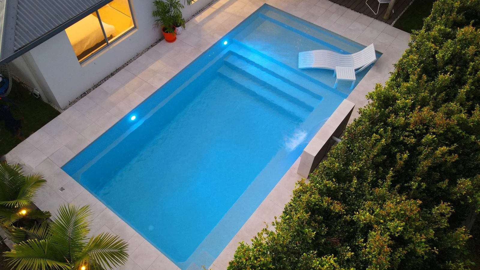 Rectangular fibreglass pool with entry steps, wide bench and cream stone paving beside a home