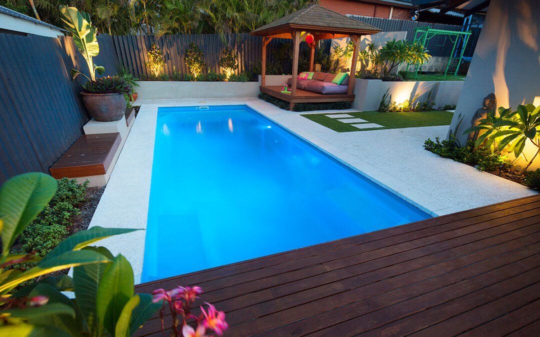 Rectangular fibreglass pool with blue water bordered by cream stone coping and a timber deck at dusk