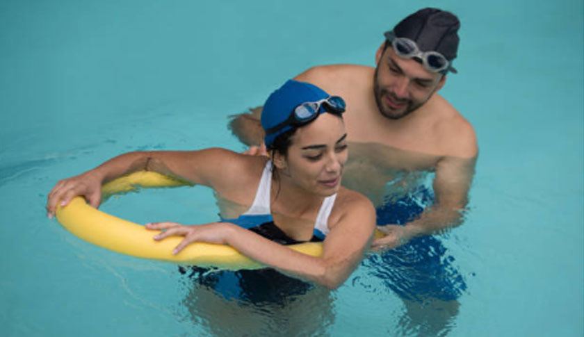 Woman using a yellow pool noodle for hydrotherapy exercises with an instructor supporting her in the water