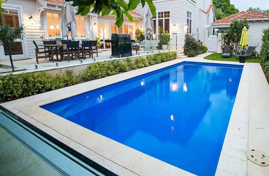 Rectangular blue fibreglass lap pool with cream stone paving beside a white house and outdoor dining area