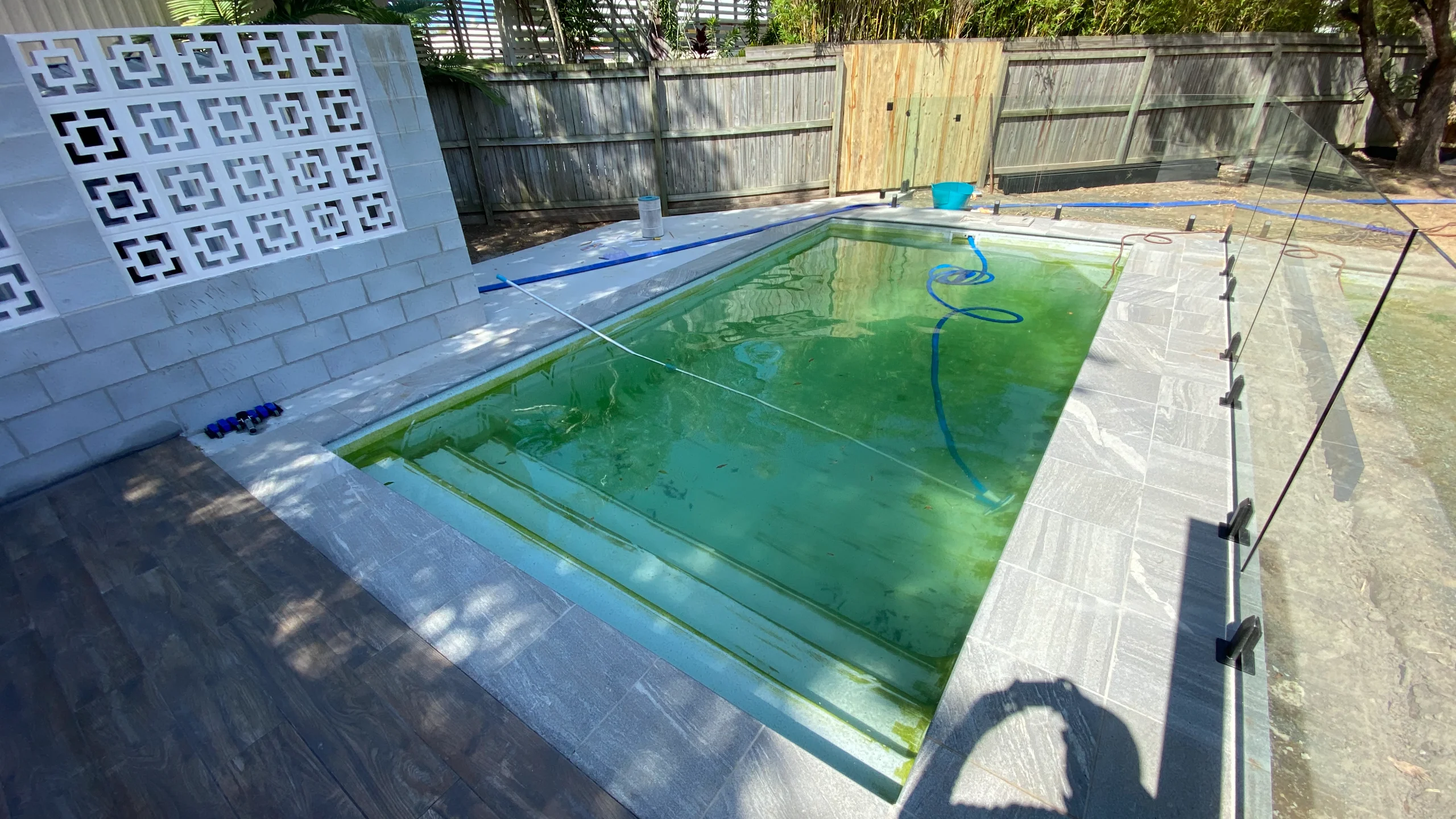 Rectangular fibreglass pool with murky green algae-affected water bordered by grey stone-look paving and glass fencing