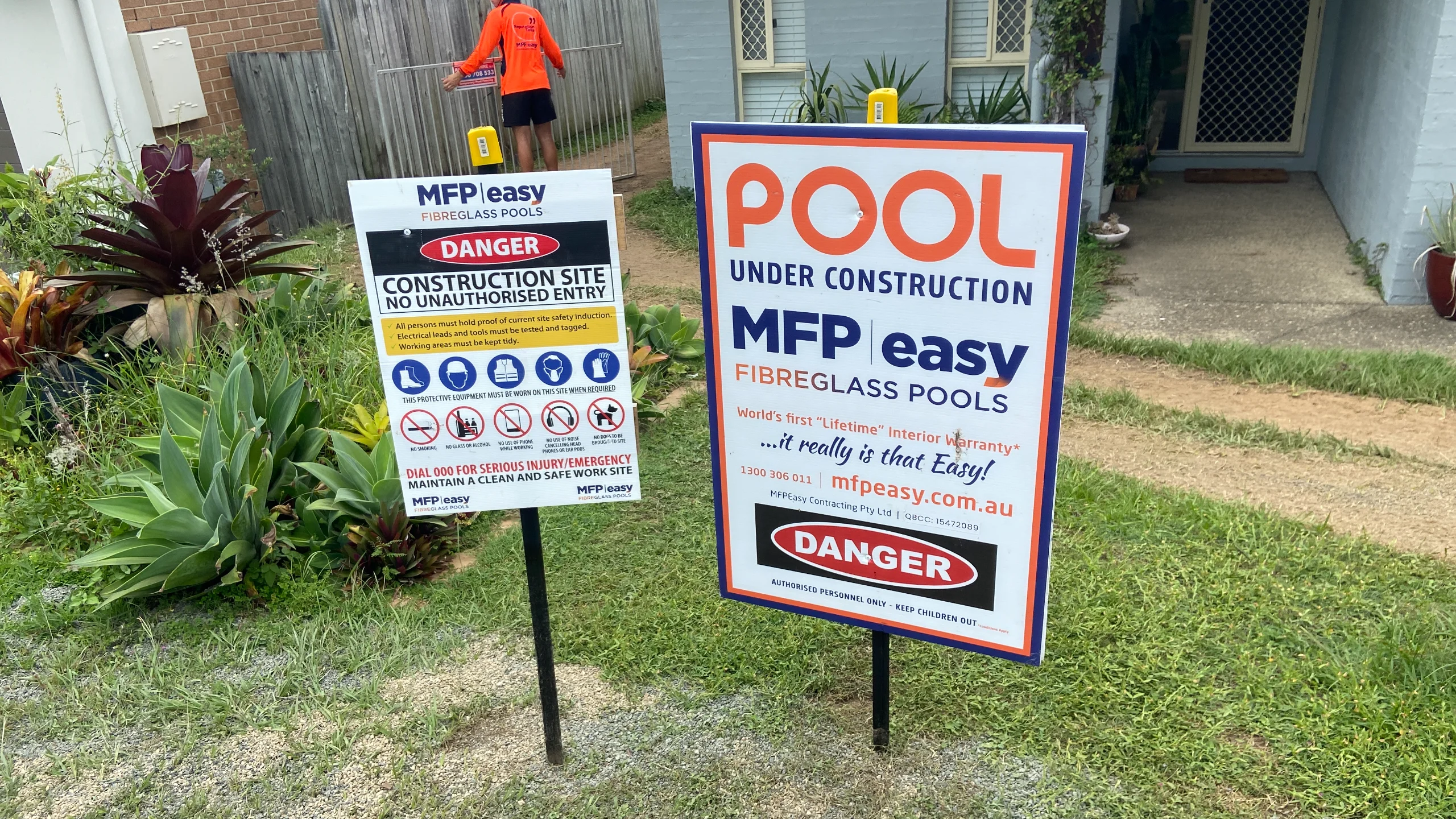 MFP Easy pool under construction danger and site safety signs staked in a grassed yard as an installer sets up temporary fencing