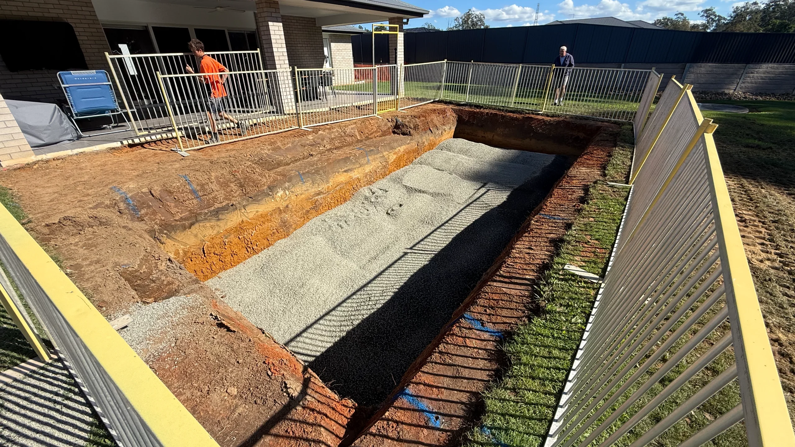 Excavated pool hole with layered soil profile and compacted gravel base behind temporary fencing in a backyard