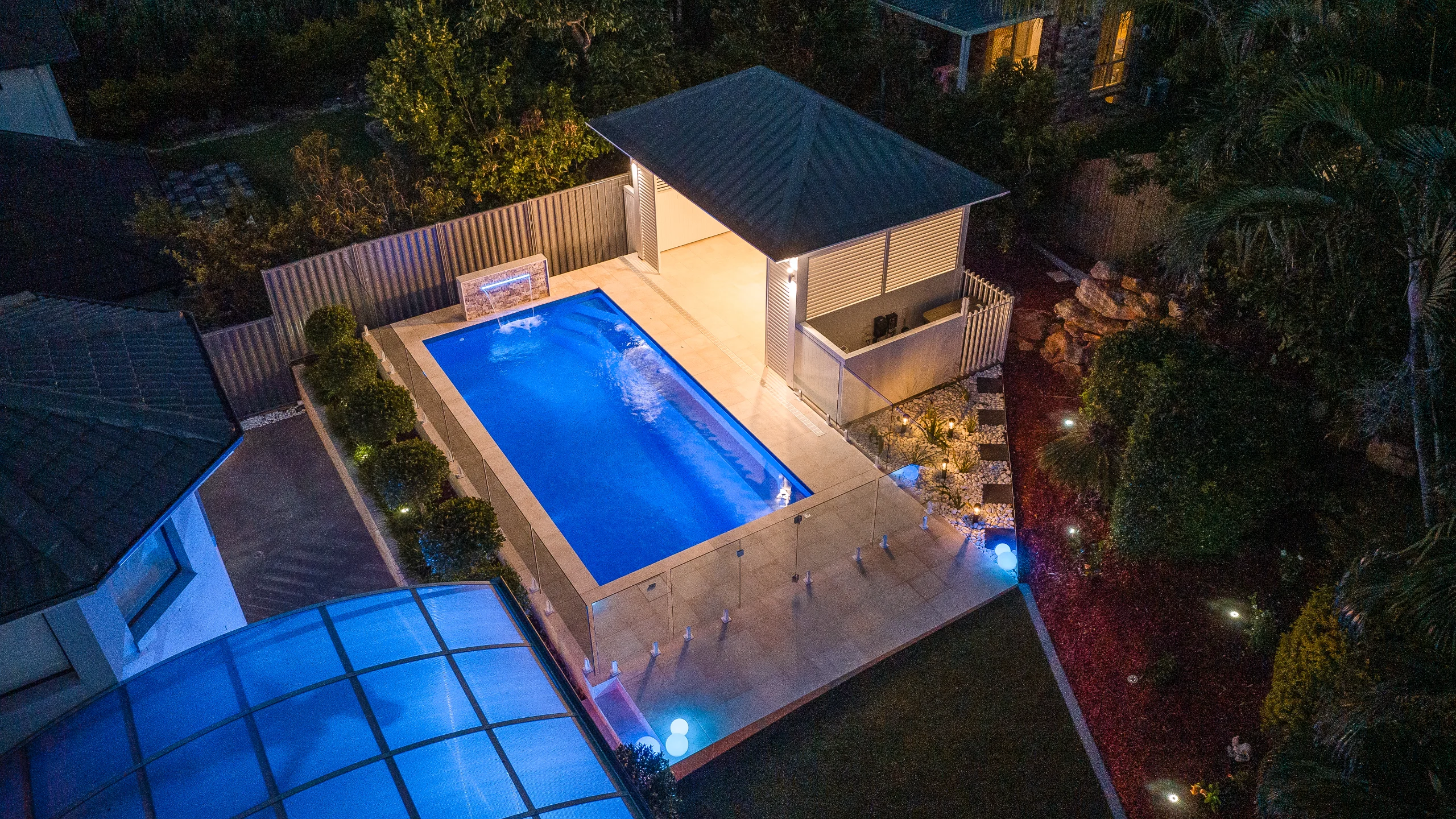 Illuminated fibreglass pool glowing blue at night with lit water feature and garden path lights