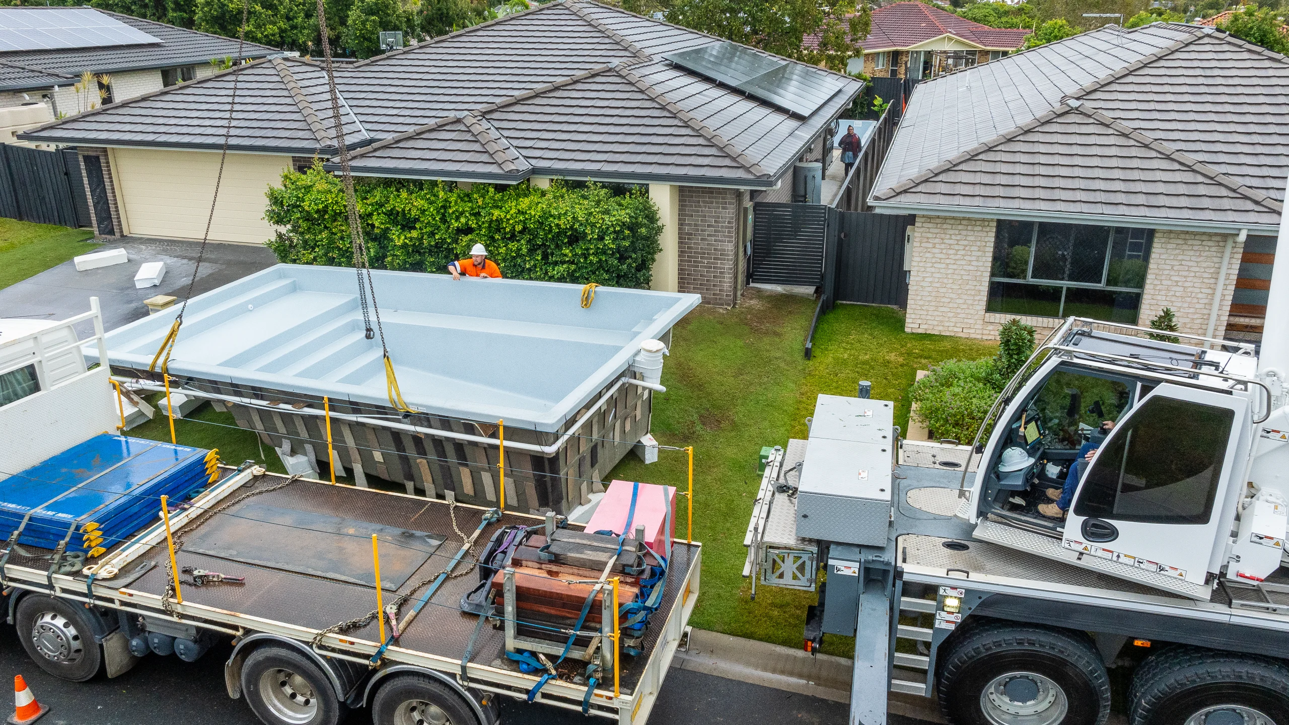 An MFP Easy installer guides a fibreglass pool shell craned over a house into a tight backyard access site