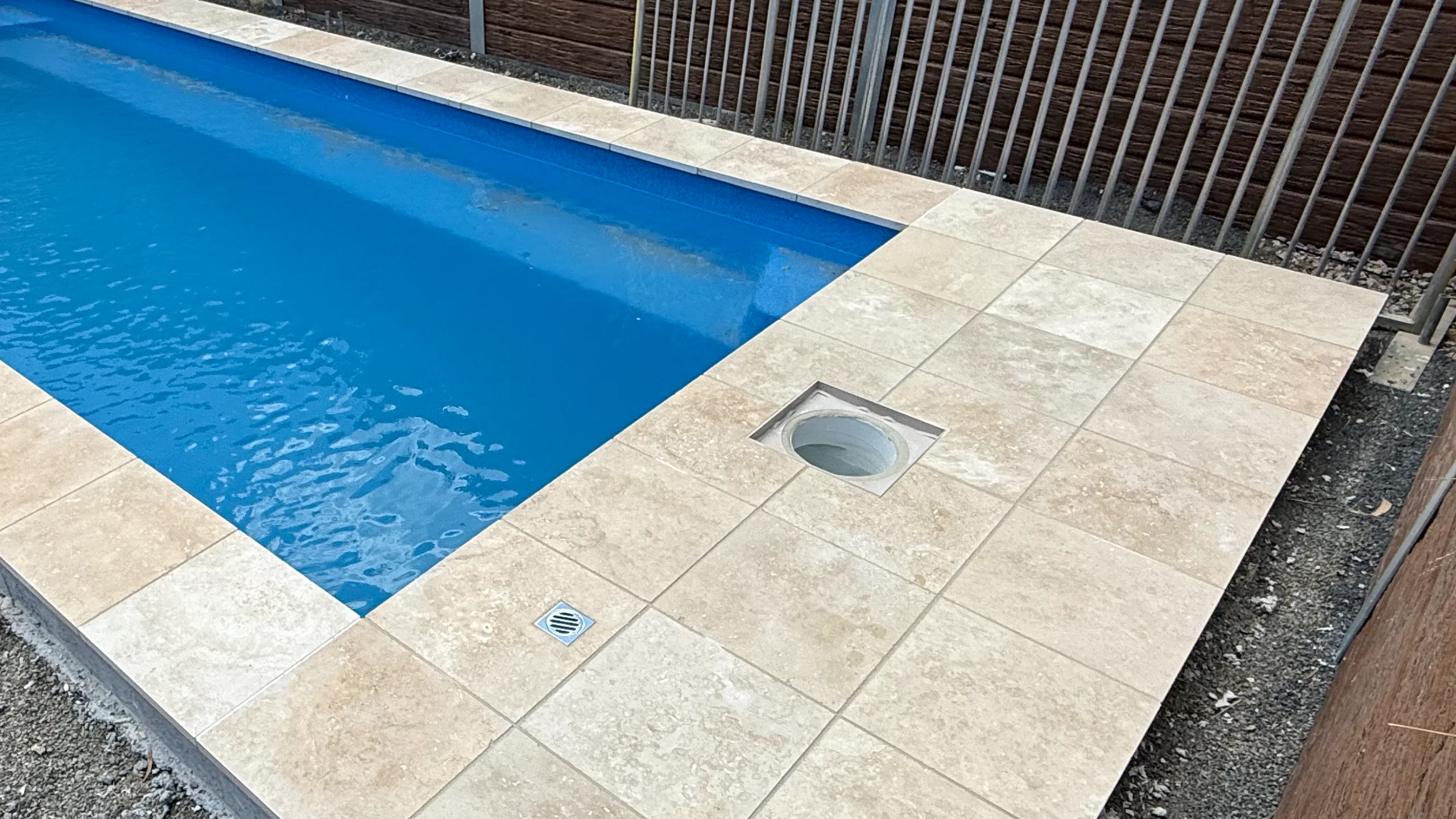 Open pool skimmer opening in cream travertine paving beside a blue fibreglass pool with a deck drain nearby