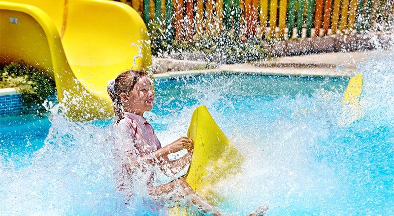 Child sliding into a bright blue pool from a yellow water slide with water splashing all around