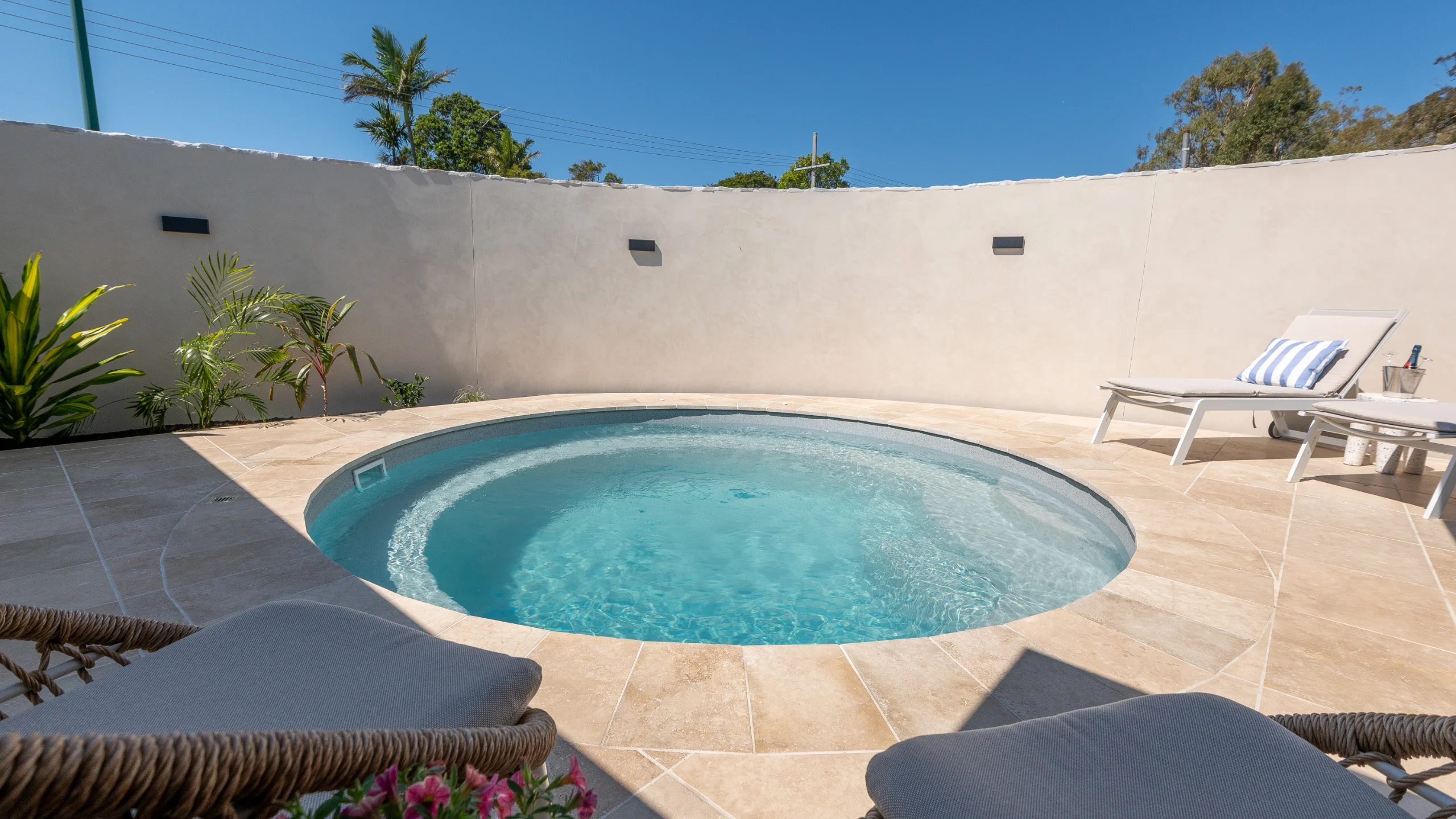 Completed round fibreglass pool with turquoise water surrounded by travertine paving, sun loungers and a rendered courtyard wall