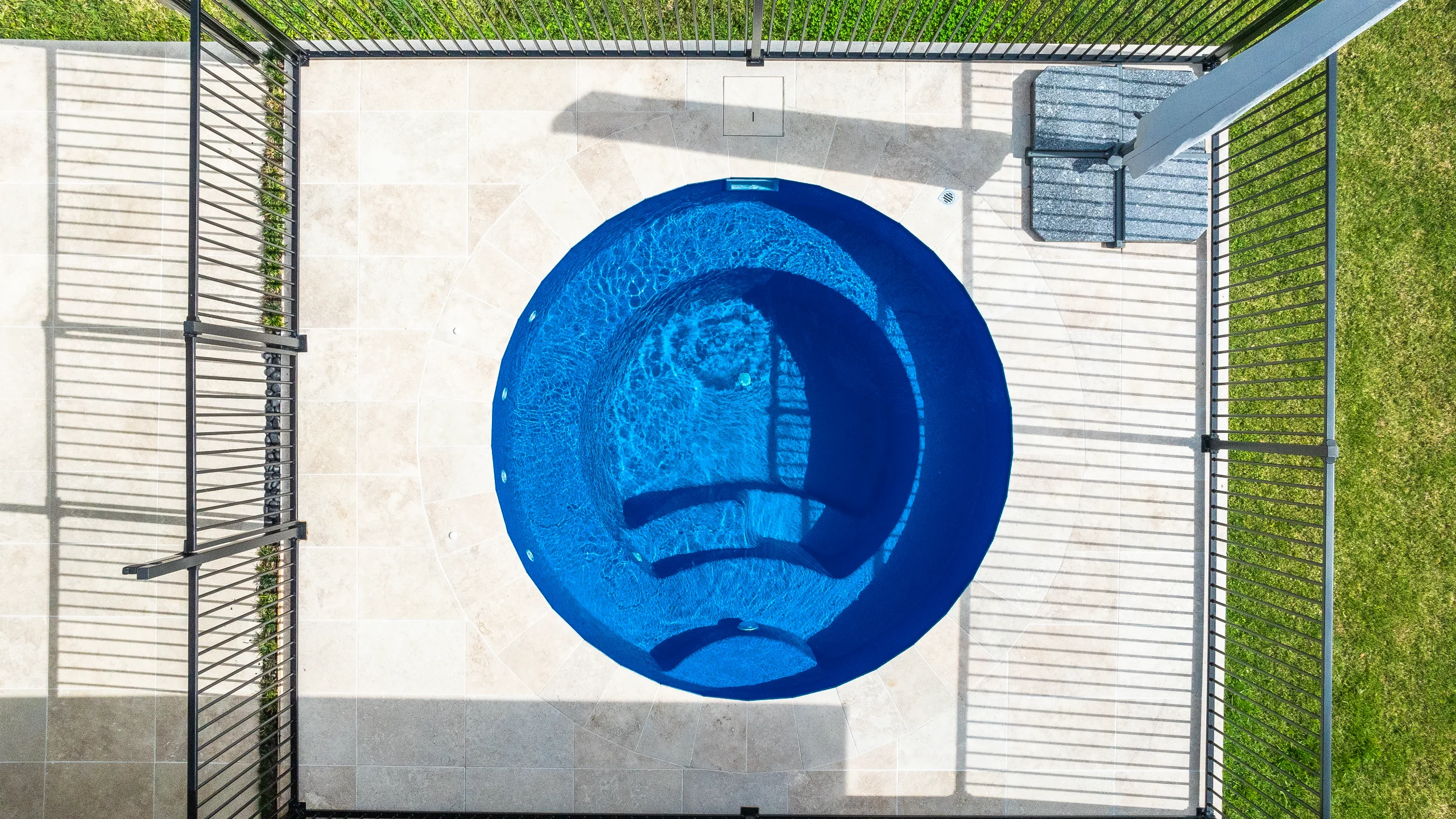 Aerial view of a round fibreglass plunge pool with built-in bench edged by cream stone pavers in a fenced yard