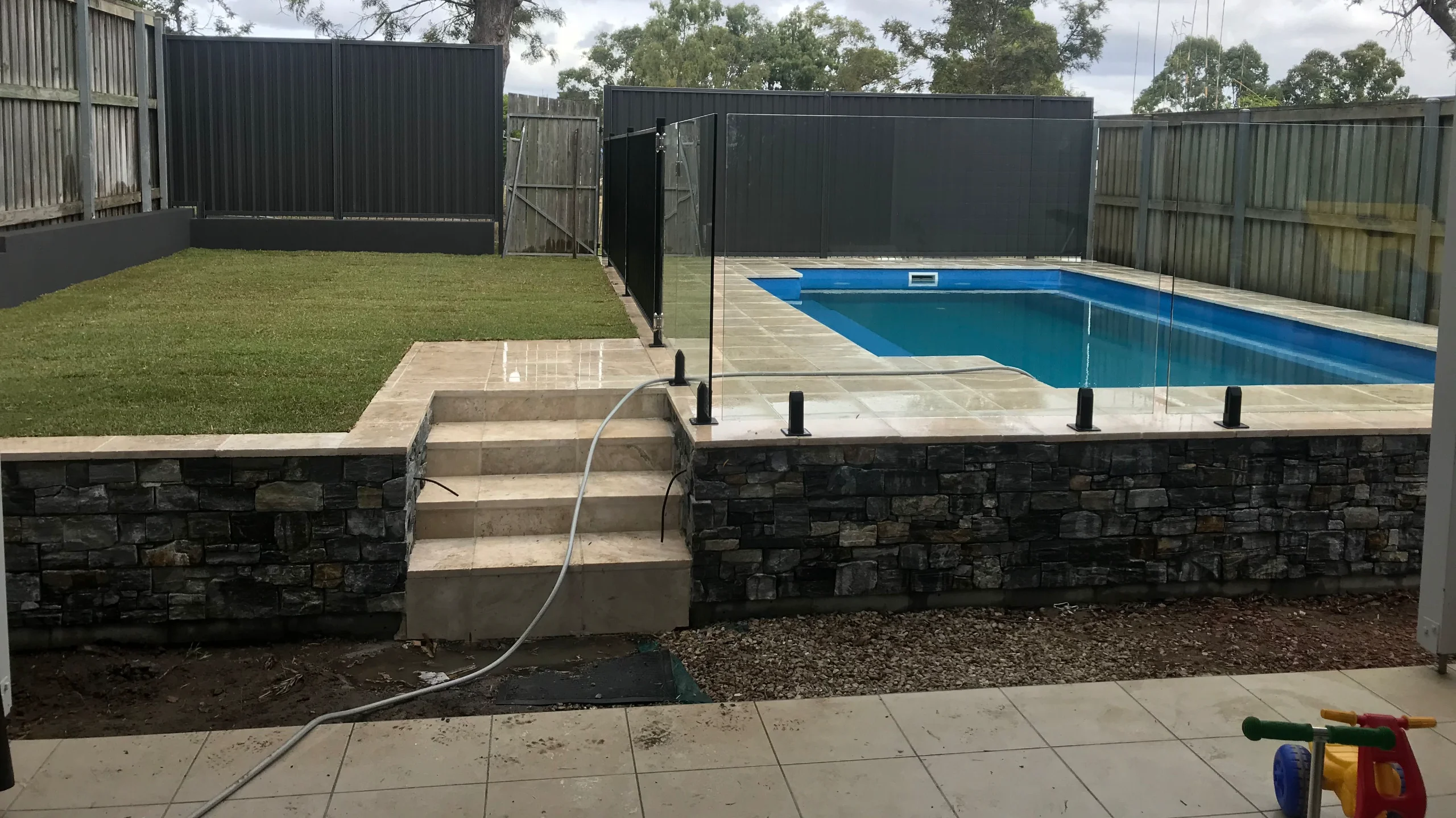 Semi-inground pool raised on a stacked stone retaining wall with tiled steps and glass fencing